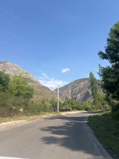 The image captures a scenic view of a winding road in the mountainous region of Brichmulla, Uzbekistan. The road, paved with asphalt, curves to the right, with grassy shoulders and some areas of exposed dirt and gravel. Power lines are strung across the clear blue sky, supported by utility poles visible in the distance.
Lush green trees and bushes line the sides of the road, creating a vibrant contrast with the arid, rocky mountains that dominate the background. The mountains themselves are rugged and imposing, with visible geological formations and sparse vegetation clinging to their slopes. The sunlight casts shadows on the road, indicating a bright, sunny day. The overall impression is one of natural beauty and a sense of remoteness, characteristic of a rural mountainous landscape. No people are visible in the image.