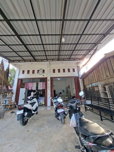The image shows an outdoor scene in front of a store in Kumbang, Indonesia. A corrugated metal roof with dark metal supports is overhead. In the foreground, two motorcycles are parked on a concrete ground. The motorcycle on the left is white, and the one on the right is black. A person is seen inside the store through an open doorway. The store appears to be a small convenience store or minimarket, with shelves stocked with various products visible behind the counter. The facade of the building has several small, dark-framed windows. To the right of the store, there is a wooden fence and a metal bench. The overall lighting suggests it is daytime.