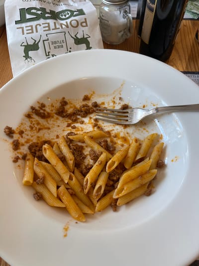 A white plate of penne pasta with a meat sauce is shown from a top-down perspective. The pasta is mostly intact, with some pieces scattered around the plate along with bits of sauce and ground meat. A fork rests on the left side of the plate, partially covered in sauce. To the left of the plate, a white paper napkin with a green logo featuring a deer and the text "FORST PONTEJEL BIER STUBE" is visible. Behind the plate and napkin, a dark glass bottle, possibly wine, is resting on a wooden surface. A small glass jar with a metal lid is also present. The scene appears to be indoors, likely a restaurant or dining setting, given the presence of the food, plate, fork, and napkin. The lighting suggests it is daytime. The location context of Cortina d'Ampezzo, Italy, is provided, hinting at the origin of the food or establishment.