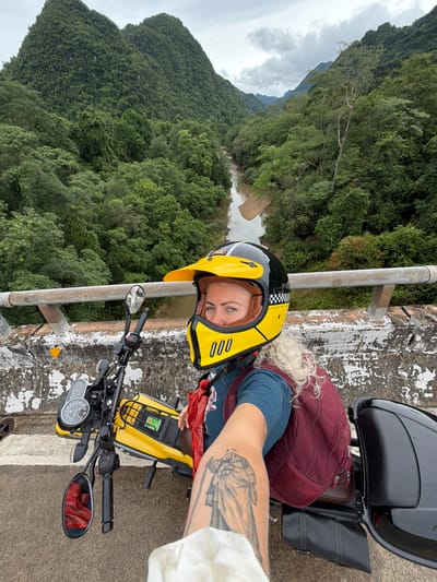 A person wearing a yellow and black motorcycle helmet is taking a selfie on a bridge. The person has blonde hair and is wearing a blue shirt and a maroon backpack. The motorcycle is yellow and black. The background shows a lush green valley with a river flowing through it and mist-covered mountains. The bridge appears to be made of concrete and has a metal railing. The area seems to be a rural mountainous region in Thượng Trạch Commune, Vietnam, known for its karst topography and natural beauty.