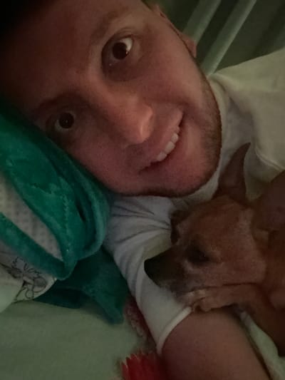 A selfie depicts a man lying down with a small brown dog resting against his chest. The man is smiling, looking towards the camera. He is wearing a white t-shirt and is partially covered by a green and white blanket. The dog's head is resting on the man's chest, facing away from the camera. The background is blurred, but appears to be bedding. There's no information to suggest this picture was taken in Vorotynsk, Russia.