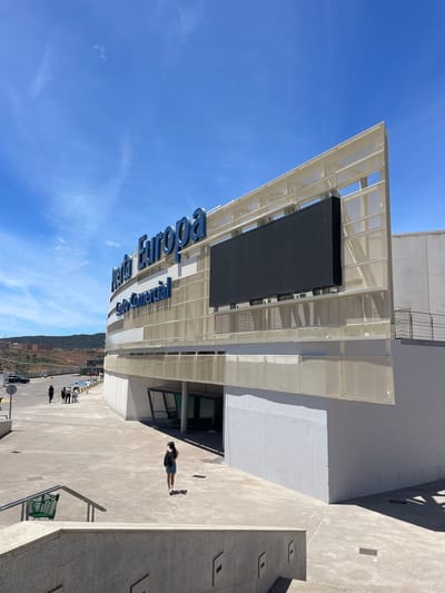 The image captures a sunny daytime scene in Algeciras, Spain. The main subject is a modern building with a large, dark digital screen mounted on its exterior. Blue, three-dimensional lettering on the building spells out "Europa Europa," indicating a location or event related to Europe. The architecture is contemporary, featuring white metal latticework and concrete elements.

In the foreground, on the paved area beside the building, a woman is standing, looking at her phone and casting a distinct shadow. She is wearing shorts and a dark top. Further in the background, several other people are visible, some standing in small groups, suggesting a public space or gathering area. A green shopping cart is also present near some railings. The setting appears to be an outdoor plaza or walkway leading up to the building, with a backdrop of a clear blue sky and some distant hills dotted with buildings, hinting at the urban edge of the town. The bright sunlight and sharp shadows indicate it is likely midday.