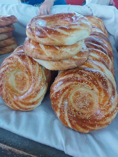 The image shows an assortment of freshly baked bread displayed on a white cloth. The bread consists of two distinct types: round, spiral-patterned flatbreads sprinkled with sesame seeds, and crescent-shaped loaves. The location is Pargos, Uzbekistan.