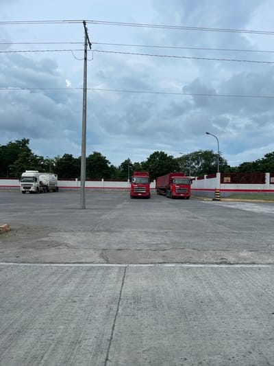 The image depicts a street scene in Santa Cruz, Philippines. On the left, power lines and a utility pole stretch upwards against a cloudy sky. Three trucks are parked along the right side of the street near a white and red wall. Two of the trucks are red, while the third one is white. The foreground consists of a concrete pavement.