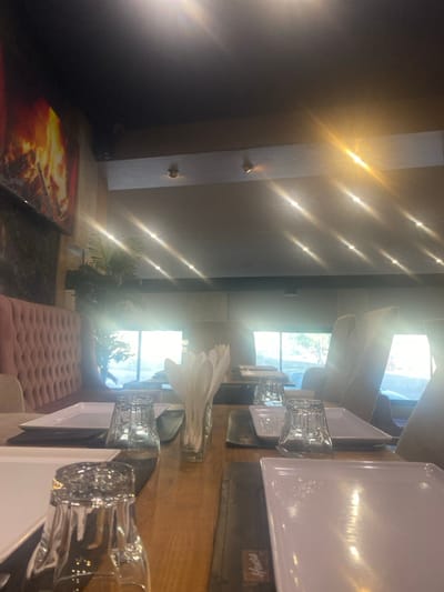 This image depicts an unoccupied restaurant interior located in Monastir, Tunisia. The scene is dominated by a long, light-brown wooden table running from the foreground to the midground. Multiple place settings are visible, each consisting of a rectangular white plate and an inverted clear drinking glass, set on dark placemats. A clear glass container holds white, feather-shaped paper napkins centrally on the table.
To the left, a light pink, tufted velvet booth runs along a wall. Above the booth, a large rectangular display or artwork shows a vibrant image of burning logs. A potted green plant with broad leaves is situated further back on the left, near windows.
On the right side of the room, several tall-backed, light-colored upholstered chairs are arranged around tables, facing large windows that span the background. These windows admit strong natural light, creating prominent lens flares across the upper half of the image and obscuring the outdoor view. The ceiling is dark, featuring recessed spotlights and a horizontal beam with additional light fixtures. No individuals are present in the scene.