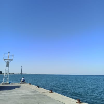 This is a photograph taken at Limani Litochorou, Greece, showing a concrete pier extending into a calm, blue sea under a clear, bright sky. On the left side of the pier, a white metal structure, possibly a navigational aid or lighthouse, stands tall. Adjacent to it, a blue bucket and fishing rods are visible. Further down the pier, a person is seated in a blue chair, facing the sea, holding a fishing rod. The pier is equipped with several metal mooring posts. In the distance, a faint shoreline with trees is visible, and a small white object, possibly a boat, is seen on the horizon. The overall scene depicts a serene day at the harbor with a lone angler.