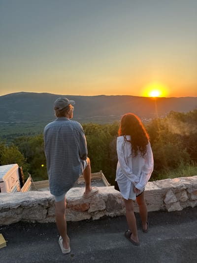 In Svinjišta, Montenegro, at sunset, two individuals stand on a stone wall overlooking a valley. The person on the left, wearing a plaid shirt, shorts, a cap, and glasses, faces away from the camera, gazing at the horizon. To the right, a person with long, curly hair, dressed in a white oversized shirt and shorts, also looks towards the sunset. The sun is a bright orb on the horizon, casting a warm glow over the landscape of rolling hills and vegetation. A construction site with a white truck is partially visible to the left of the frame. The sky transitions from a pale blue at the top to vibrant orange and yellow near the sun.