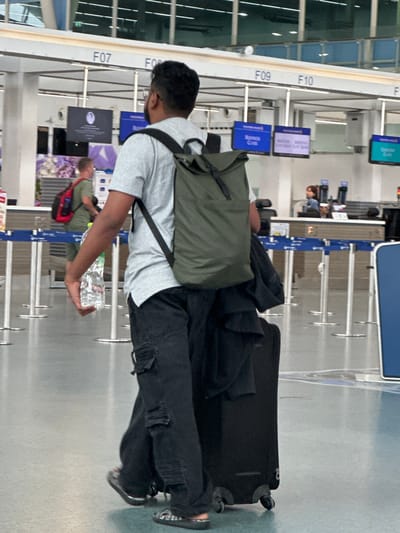An adult male is walking through an airport terminal in Tambon Mai Khao, Thailand. He is wearing a light grey t-shirt, black cargo pants, and dark slip-on shoes. He has a black backpack with olive green straps on his back and is carrying a clear plastic bottle of water in his right hand. He is also pulling a black rolling suitcase. The man is walking away from the camera, towards a row of check-in counters with signs indicating gates F07, F09, and F10. Several other individuals are visible in the background, including a person at a check-in desk and another person with a red backpack walking in the distance. The terminal has a polished floor and a light-colored ceiling with exposed infrastructure.