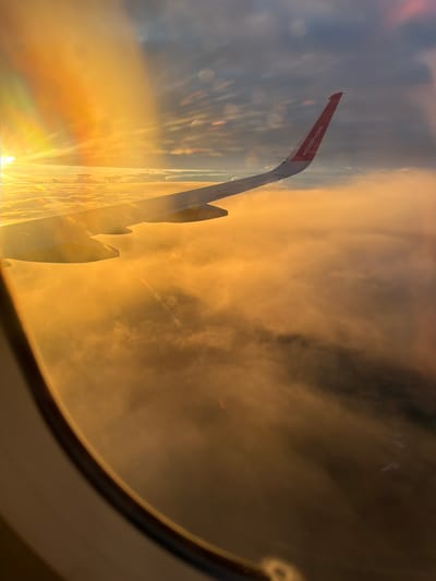 This is an aerial photograph of clouds from the perspective of an airplane window, likely taken during sunrise or sunset due to the warm, golden light. The sun's rays are visible, creating a lens flare effect. An airplane wing, with the Wizz Air logo visible on its tip, extends into the frame from the right side. The clouds below appear dense and illuminated by the sun. The bottom left of the image shows a portion of the airplane's oval-shaped window frame. There is no information in the image that specifically indicates the location is Czerwony Kościół, Poland.