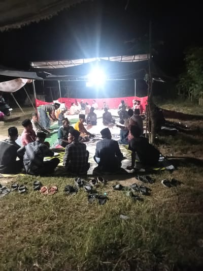 In Matangkareung, Indonesia, an outdoor gathering is taking place at night. A group of approximately 25 individuals, predominantly male, are seated on the ground, facing a central area under a temporary shelter. The shelter appears to be constructed from fabric stretched over a frame, illuminated by a bright overhead light source, casting strong shadows. The attendees are dressed in casual attire, many wearing caps or skullcaps. The ground is covered with grass and mats, with numerous pairs of sandals scattered around the perimeter. In the background, trees and vegetation are visible, suggesting a rural or semi-rural setting. The overall atmosphere suggests a community event or religious observance.