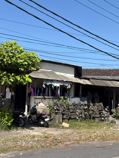 The image depicts a roadside scene in Sukawati, Indonesia, featuring a small shop or stall selling carved stone statues. The shop has a corrugated metal roof extending over an open-air display area. In front of the shop, a dense collection of intricately carved stone statues, many appearing to be of mythical creatures or deities, are arranged on the ground. Some of these statues are on pedestals. A motorcycle is parked to the left of the statues, and another motorcycle is parked in front of the shop. Above the shop, laundry hangs on a clothesline. A large, leafy green tree with bright green foliage is positioned to the left of the frame, partially obscuring the shop's entrance. Power lines are visible crisscrossing the bright blue sky overhead. The ground in front of the shop is a mixture of dirt, gravel, and sparse dry grass, with some litter visible on the roadside. The overall impression is a typical commercial streetscape in a tropical location.