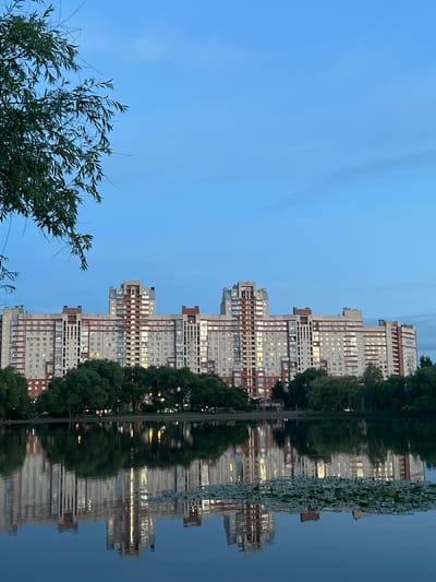 This media file depicts a reflected view of a residential building and trees in water. The building is tall and modern, with many windows, and is a pale color with reddish-brown accents. Trees, lush and green, are visible in front of the building and also reflected in the water. The scene is set against a clear, pale blue sky, suggesting it is either early morning or late evening. The water's surface is slightly disturbed, creating a rippled reflection of the building and trees. The overall mood is tranquil and serene. There is no visible text in the image. The location is Rahya, Russia.