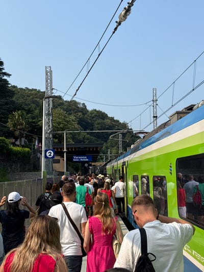 The image shows a crowd of people waiting on a train platform in Varenna, Italy. A bright green and blue train is stopped at the station. The people are standing close together, suggesting they are waiting to board the train or have just disembarked. Some individuals are looking towards the train, while others are interacting with each other. The scene appears to be set on a sunny day, with a clear blue sky visible. The background features lush green trees and foliage, indicating a natural, possibly hilly, landscape surrounding the station.
Notable details include the train's vibrant color scheme and the presence of overhead power lines, common for electric trains. A sign on the platform reads "Varenna - Esino - Perledo," indicating the station's location and the destinations served by the train. Another sign displays the number "2" in a blue square, likely representing the platform number. The overall atmosphere seems to be one of travel and transit, with people appearing to be either arriving or departing for their destinations. The presence of luggage and the way people are dressed suggest it might be a tourist season.