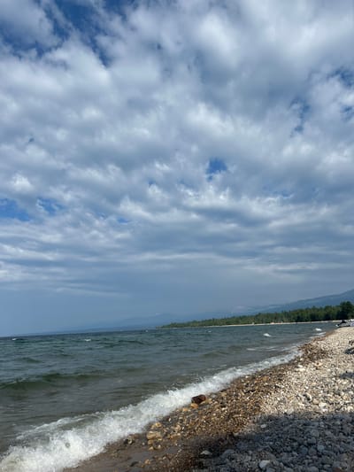 The image shows a wide, pebble-strewn beach along a large body of water. The water is a dark, choppy green with white-capped waves rolling onto the shore. The sky is filled with a dramatic arrangement of grey and white clouds, with patches of blue sky visible between them. In the distance, the shoreline is lined with a dense forest of green trees, beyond which faint, hazy mountains can be seen. A few scattered tents and vehicles are visible along the tree line. The overall impression is of a natural, somewhat wild landscape. The exact location in Tolbazikha, Russia, is not specified in the visual information.