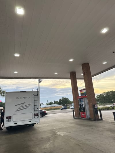 A white Chateau RV is parked at a RaceTrac gas station in Palm Bay, Florida. The back of the RV, including its ladder, is visible. A man is standing behind the RV. A car is visible in the background. The gas station has brick pillars and a price display. The sky is visible in the background.