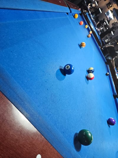 A low-angle, oblique shot captures the blue-felt surface of a pool table within an indoor billiards hall in Arnhem, Netherlands. The foreground features the polished, reddish-brown wooden rail of the table. Numerous pool balls are scattered across the felt. A green solid ball (6-ball) rests near the bottom left, with a purple solid ball (4-ball) positioned further right. Towards the center-left, a blue solid 2-ball is prominent. Further right, a red-and-white striped 9-ball and a blue-and-yellow striped 10-ball are in close proximity. A black 8-ball is visible closer to the mid-right side rail. A cluster of balls is seen towards the upper right side of the table, including a yellow solid 1-ball, an orange solid 5-ball, a red solid 7-ball, a yellow-striped 9-ball, an orange-striped 13-ball, and a brown-and-white striped 15-ball. The blue felt exhibits minor dark flecks and slight wear. In the dimly lit background, several other pool tables are visible, each illuminated by overhead lamps with black shades. Multiple individuals, adults, are present in the background, some appearing to be playing at other tables, while others observe, indicating a recreational social setting.