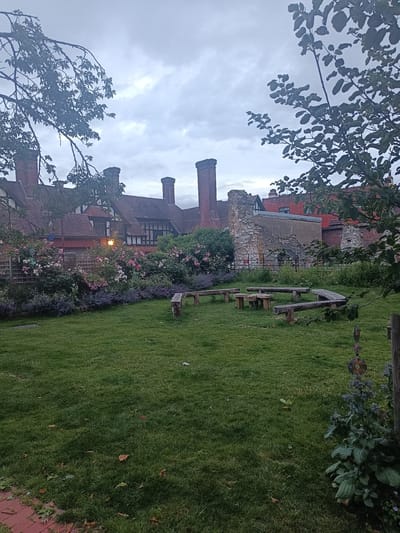 The image depicts a garden area with a view of a historic building in the background. The foreground consists of a green lawn with several wooden benches arranged in a semi-circular fashion, accompanied by a few low wooden tables. Lush greenery, including flowering bushes with pink and purple blooms, borders the lawn. In the background, a large building with red-tiled roofs, multiple chimneys, and timber-framed architectural elements is visible. Part of the building appears to be in a state of ruin, with exposed stone walls. The sky is overcast, suggesting a cloudy day. The scene is serene and appears to be a public or communal garden space within Arun, United Kingdom, possibly a historical site due to the architecture and ruins. No people are present in the image.