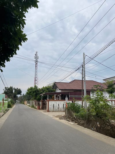 A photograph taken from the perspective of standing on an asphalt road in the city of Tanahterban, Indonesia, shows a street lined with houses and a telecommunications tower in the distance. The sky is overcast with a soft gray light. On the left side of the road, a green gate and part of a house are visible, with lush green foliage dominating the foreground. The right side of the road features a white house with a reddish-brown tiled roof, a white fence, and more vegetation. Several power lines crisscross the sky, supported by a utility pole in the mid-ground. The telecommunications tower, a prominent structure, stands tall in the background, with its red and white frame and antennas clearly discernible. A small section of a bamboo fence runs along the side of the road, overgrown with green plants. A person is partially visible in the background, near the white house.