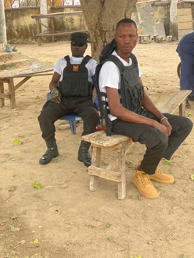 Two adult males are observed seated outdoors in Gidan Kado, Nigeria, under a large tree and adjacent to a light-colored building. The ground is composed of reddish-brown dirt, sparsely covered with fallen dry leaves.
The male on the left is seated on a blue plastic chair. He wears a black wide-brimmed hat, dark sunglasses, a white short-sleeved t-shirt, a black tactical vest bearing an "NHSS" logo with a red and yellow insignia, black tactical cargo pants, and black combat boots. A camouflage-patterned glove is worn on his left hand. His posture is relaxed, with his head turned slightly to his right.
The male on the right is seated on a crude, unpainted wooden stool. He wears a white short-sleeved t-shirt, a black tactical vest without a visible insignia, black tactical cargo pants, and tan work boots. A black backpack is slung over his right shoulder. His gaze is directed forward.
In the background, several rudimentary wooden benches or tables are visible. A light-colored building with some red trim is partially visible behind the subject on the left. The scene is illuminated by ambient daylight.