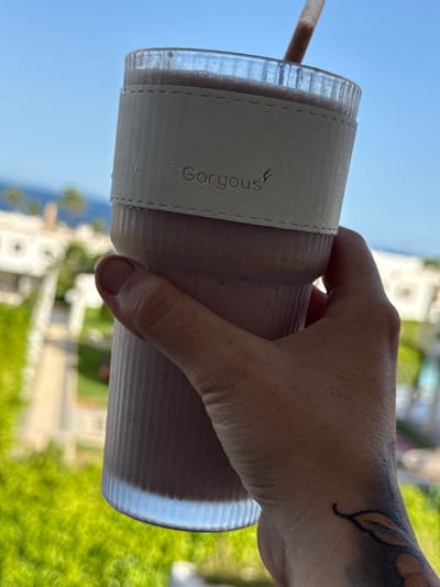 A close-up, vertical, slightly upward shot shows a hand holding a ribbed, translucent cup filled with a pinkish-purple smoothie. The cup has a white band around it with the word "Gorgous" embossed in a cursive font and a small leaf logo. A straw extends from the top of the smoothie. The background is blurred, showing a bright blue sky above, a body of water, some palm trees, and buildings, suggesting an outdoor setting, likely a resort or coastal area. The hand exhibits a tattoo of a leaf on the wrist. The overall lighting indicates a sunny day. The image appears to have been taken in Aguadulce, Spain, given the context of a resort environment with Mediterranean-style architecture and lush greenery.