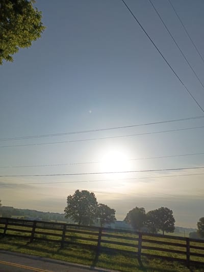 This image depicts a rural landscape at sunrise. The foreground is dominated by a clear blue sky with several parallel power lines stretching across it. To the right, a wooden fence borders a grassy field. In the distance, trees are silhouetted against the bright, hazy sky, suggesting a foggy or misty morning. The sun is low on the horizon, casting a bright glow and creating lens flare. The overall mood is serene and peaceful, evoking a sense of early morning tranquility. There is no visible text in the image.