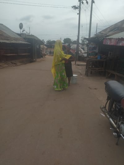 Two women are standing on a dusty road in Junction, Nigeria, at coordinates 8.958, 7.551. One of the women is wearing a yellow hijab and a long green skirt with a floral pattern, while the other is wearing a black dress. There is a white bucket next to them. There are buildings on both sides of the street, with some appearing to be shops. A motorcycle is parked on the right side of the street. There are power lines and poles above the street. The sky is overcast and there are some trees in the distance.