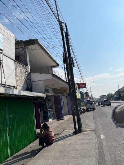 The image captures a street scene in the city of Gedang, Indonesia, bathed in bright sunlight under a clear blue sky with scattered clouds. A person, dressed in a reddish-brown top and dark pants, is seen squatting on a concrete sidewalk, their head bowed and hands near their face, appearing contemplative or distressed. To their left is a closed green metal roller door on a building.

The buildings lining the street are modest, with some showing signs of wear. One building displays signage for "TOKO ASIA ELEKTRONIK" and "JUAL KACA ALUMINIUM," indicating commercial establishments. Power lines crisscross overhead, supported by tall poles.

The street itself is paved and carries traffic, including cars, motorcycles, and a truck. The sidewalk runs parallel to the road, separated by a curb. The overall environment suggests a typical urban or semi-urban setting in Indonesia, likely during daylight hours. The perspective is from a slightly elevated position, looking down the street and towards the traffic.
