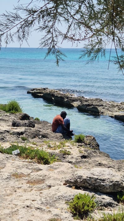 A person sits on a rocky outcrop overlooking a body of clear turquoise water. The person is facing away from the camera and appears to be wearing an orange shirt and blue shorts. They are crouched down, with their legs drawn up. The rocks are rough and textured, with some sparse green vegetation growing between them. The water is calm and reflects the bright sky, suggesting a sunny day. The scene is likely a coastal area, given the rocky terrain and the sea. The image is taken from a slightly elevated perspective, with the branches of a tree framing the left side of the frame. The overall mood is peaceful and serene. No visible text is present in the image.