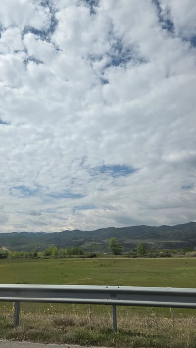 The image shows a landscape taken from what appears to be the side of a road, possibly in Strumyani, Bulgaria. A guardrail is prominently visible on the right. Beyond the guardrail is a field and a mountain range. The sky is predominantly covered with white clouds, interspersed with patches of blue.