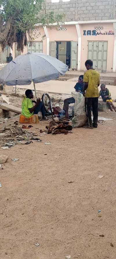 The image depicts an outdoor scene in Rijiyar Zaki, Nigeria. An adult male wearing a green shirt sits on a small orange crate beneath a large, light-colored umbrella, apparently repairing a bicycle tire. Near him, another adult male sits on the ground next to a large plastic sack. A third adult male, wearing a yellow shirt, stands near the bag. To the right of the standing man, a child sits on a concrete structure. Discarded materials and litter are scattered on the ground around the group. Behind them is a storefront with the sign "IASProvision". The foreground is a dirt area with scattered debris.