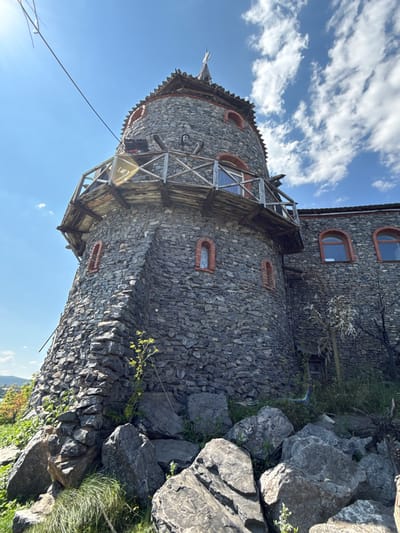 The image shows a stone tower, possibly part of a building or structure, with a wooden balcony on its upper section. The tower is constructed from irregular gray stones and features small arched windows with red brick surrounds. The surrounding area appears to be natural, with large rocks and vegetation at the base of the tower. The sky is blue with scattered clouds, and sunlight is visible. There are no people or active events depicted in the image. The tower's design suggests a rustic or historical aesthetic.