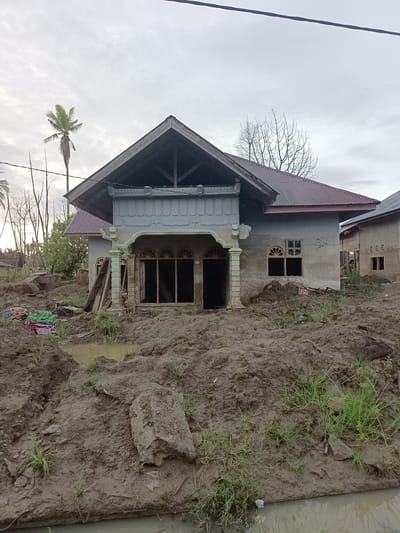 The image depicts the aftermath of a disaster, likely a mudslide or flood, in the city of Meunasah Raya, Indonesia. In the foreground, there is a significant accumulation of thick, dark mud, with pools of murky water interspersed throughout. Patches of green grass are visible pushing through the mud, indicating some attempt at recovery or natural regrowth. Several large rocks are embedded in the mud.

Dominating the mid-ground is a partially constructed or damaged house. Its roof is made of corrugated metal, some of which appears to be a reddish-brown color, while other sections are a dark grey, suggesting different materials or weathering. The front of the house has a covered porch area with decorative pillars. The windows and doorways are open and dark, indicating the absence of glass or doors. A wooden ladder leans against the side of the house near the porch. To the right of this main structure, another building with a similar roofline and concrete walls is visible, suggesting a residential area. In the background, several bare tree branches reach towards an overcast sky. A single palm tree with green fronds stands to the left of the main house. Piles of colorful fabrics or tarps are scattered on the mud to the left of the house. The overall scene conveys a sense of devastation and ongoing reconstruction or clean-up efforts.