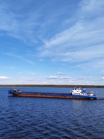 A long blue and brown barge moves across a body of water under a partly cloudy blue sky. The barge has a blue tugboat section at the front and the back. The body of water extends to the horizon where there is a thin line of trees. The location is Gorushka, Russia.