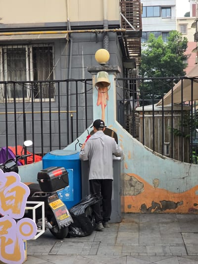 In Gongshu District, China, a person wearing a grey hooded jacket and a black baseball cap is standing with their back to the camera, facing a light blue utility container and a grey utility box. They are in the process of discarding a black plastic bag into the utility box. To the left of the person, a black motorcycle is parked, its rear tire and license plate visible. The license plate displays the number "064560" and some Chinese characters. In the background, a weathered, light blue and orange painted wall with peeling paint forms a curved structure. Beyond the wall, buildings with windows and trees are visible. A yellow gas pipe runs horizontally above a barred window on a grey building to the left.