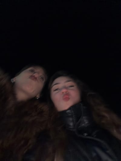 This close-up image features two young women posing in a dimly lit environment, likely at night in Lampertheim, France. The primary subject, positioned in the upper right, has long, dark, wavy hair and wears a dark jacket, possibly leather. Her face is illuminated by a flash, highlighting her playful expression with pursed lips (a "kissing face") and wide eyes. Below and slightly to her left, the second woman is partially visible, also with long dark hair and wearing an earring, mimicking the same "kissing face" expression. The background is completely black, offering no discernible details of the surroundings and drawing full attention to the subjects. The image is slightly blurry and tilted diagonally, suggesting a casual, spontaneous selfie or snapshot. The overall mood is lighthearted and friendly. No text is visible within the image.