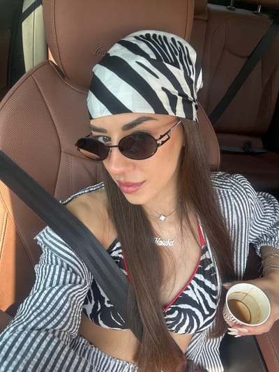 A woman with dark hair, wearing a zebra-print bandana and sunglasses, is seated in the driver's seat of a car. She is wearing a zebra-print bikini top and a striped button-up shirt, with a silver necklace and bracelets. She holds a paper cup of coffee in her right hand. The car interior appears to be leather, with a brown headrest visible. The scene is lit by sunlight streaming in from the left. The location is specified as Neo Souli, Greece.