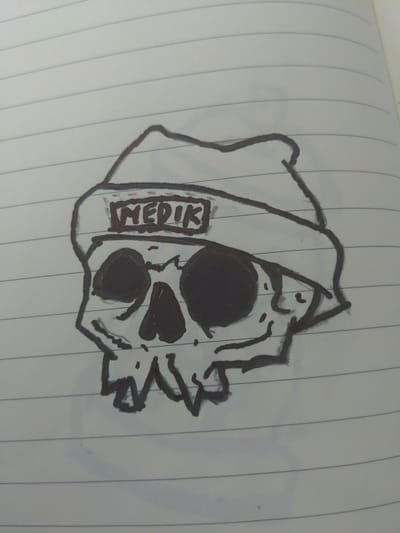 The image is a close-up, slightly angled, overhead shot of a hand-drawn sketch on lined notebook paper. The drawing depicts a stylized skull wearing a hooded robe, with the hood pulled up to cover the top of the skull. The skull has large, dark circles for eye sockets and a smaller, darker shape for a nose cavity. A row of sharp, jagged lines suggests teeth. To the left of the skull, within the folds of the robe, is a rectangular label with the word "MEDIK" written in uppercase letters. The drawing is executed with thick, dark lines, likely with a marker. The background is the white lined paper, with faint blue lines running vertically across the image. The overall impression is a cartoonish or symbolic representation, possibly related to themes of medicine or death, rendered in a simple, bold style. There are no overt indications of time of day, weather, or specific emotions beyond the implied symbolism of the skull. The location context is Kamen, Germany, but there are no visual cues within the image itself to support this.