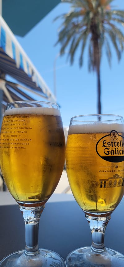 Two chilled glasses of Estrella Galicia beer are prominently displayed, holding a golden-yellow liquid topped with a foamy head, suggesting freshness. Condensation is visible on the sides of both glasses, indicating they are cold. The top glass clearly features the "Estrella Galicia" logo, a star, and the words "CERVECEROS DESDE" (Brewers since). The lower glass also has text printed along its side, which reads: "LA PERFECCIÓN OBTENIDA EN LA ELABORACIÓN DE ESTA CERVEZA, ES FRUTO DE LA EXPERIENCIA TRANSMITIDA A TRAVÉS DE LOS AÑOS Y CONSTANTEMENTE MEJORADA POR CINCO GENERACIONES DE LA FAMILIA RIVERA."

The beers are resting on a dark, flat surface, likely a table. In the background, a vibrant blue sky dominates, indicating bright, clear weather during the daytime. A blurred palm tree is visible in the upper left, adding to a tropical or warm-weather ambiance. Further in the background, a white and blue striped structure, possibly a canopy, awning, or part of a boat, is out of focus. The scene evokes a relaxed moment, enjoying refreshing drinks in an outdoor setting, consistent with the given location of Premià de Mar, Spain.
