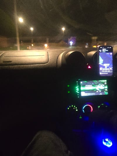 The image depicts the interior of a vehicle at night, positioned in Elmbridge, United Kingdom. The scene is viewed from an occupant's perspective. In the immediate foreground, a portion of a human leg, covered in grey fabric, is visible. The vehicle's dark grey dashboard occupies the mid-ground. An illuminated digital display screen in the center console shows a green-themed interface, possibly a media player. Below this, circular climate control dials glow green, red, and blue. A red triangular hazard light button is illuminated. To the right, a smartphone is mounted on the dashboard, actively displaying a navigation application. The phone screen shows a map with a purple and blue color scheme, indicating a route from "Elmbridge" towards "London." A prominent blue light source emanates from the lower right console area, likely from an accessory. Through the windshield, multiple streetlights are visible, illuminating the exterior environment, which consists of a road and indistinct background structures under dark conditions.
