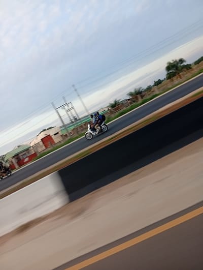 This tilted photograph depicts a dynamic road scene in Uke, Nigeria. A white underbone-style scooter, carrying three individuals, is prominently featured in the mid-ground, moving from right to left across the frame. The driver wears a light-colored shirt and dark pants, with two male passengers behind him, one in a blue shirt and the other in a dark shirt. In the left foreground, a second, partially visible dark motorcycle is also in motion, traveling in the same direction. The asphalt road consists of multiple lanes, bordered by a light-colored concrete barrier and a darker inner section, with a distinct yellow line marking the far right lane. The background exhibits significant motion blur, showcasing utility poles, power lines, and several buildings, notably structures with green roofs and prominent red doors. Sparse vegetation and open land stretch into the distance under a pale, overcast sky. The image captures a moment of vehicular transit on a major thoroughfare.