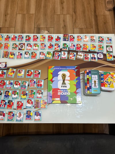 The image displays a collection of FIFA World Cup 2026 stickers and related merchandise. A sticker album and two tin boxes, one blue and one green, are prominently featured on a white surface. The tin boxes contain various stickers, including one with the text "WE ARE ONE FIFA" and another showcasing a soccer player. Scattered around these items are numerous individual stickers, each featuring a portrait of a soccer player with their name and country. These stickers are arranged in rows, suggesting a hobby or collection. The background consists of a wooden floor. The scene is indoors, and there are no people or animals visible in the immediate vicinity. There is no indication of the city of Prisovo, Bulgaria in this image.