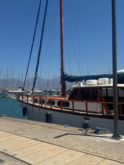 This image captures a docked sailboat with a clear blue sky overhead. The sailboat features a prominent mast and sails, and its wooden railings and white hull are visible. Several other sailboats are lined up behind it, their masts creating a dense forest against the sky. The boat is moored at a concrete dock, with ropes and fenders secured. In the background, mountains are visible in the distance, suggesting a coastal or island location. The bright sunlight indicates it is daytime, and the clear sky points to fair weather. The scene is serene, with no people visible. The overall impression is one of a peaceful harbor on a sunny day.