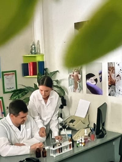 In a laboratory setting, two individuals, a man and a woman, are dressed in white lab coats. The man is seated at a desk, looking down at the test tubes in front of him. The woman stands behind him, leaning over the desk, and appears to be assisting or instructing him. On the desk, there are various laboratory equipment and supplies, including a microscope, a beige-colored machine with a keypad, bottles, and a rack of test tubes filled with liquids. The walls of the lab are painted white, and a green shelf holding books and a small decorative item is mounted on the wall. Three framed photographs are also visible on the wall, depicting close-ups of faces and hands, possibly related to scientific or medical procedures. The overall environment suggests a controlled research or educational space within a laboratory. The location is not discernible from the provided image.