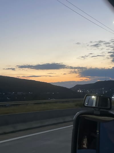 This is a photograph taken from a moving vehicle, looking towards a sunset over a valley. The sky is a gradient of soft blues and oranges, with scattered clouds. In the foreground, a side-view mirror of the vehicle is visible, reflecting the sky and interior of the car. Beyond the mirror, a highway is seen, with a dark mountain range in the middle ground. Small lights, indicative of a town or village, dot the slopes of the mountains. The location is identified as Changilari, Georgia.
