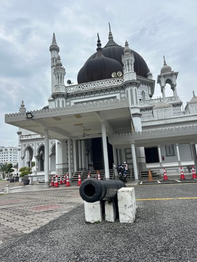 The image depicts the Masjid Zahir in Alor Setar, Malaysia, on an overcast day. In the foreground, a weathered, dark-colored cannon barrel is mounted on two white concrete blocks, positioned on a gravel surface. Behind the cannon, a paved walkway with a patterned design leads towards the mosque's entrance. Red and white traffic cones are lined up along the edge of the walkway. The mosque itself is a grand white structure with intricate architectural details, featuring multiple domes, minarets, and arched doorways. A motorcycle is parked on the steps leading to the entrance. In the background, a modern white building is visible to the left, suggesting an urban setting. The sky is heavily clouded, casting a subdued light on the scene.