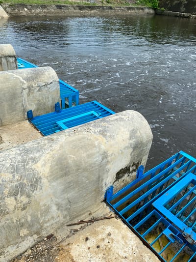 The image shows a section of an irrigation canal or water management structure. The canal is filled with dark, murky water with visible ripples and some white foam on the surface. Along the edge of the canal, there are concrete structures with rounded tops. Attached to these concrete structures are blue metal grates or screens, which appear to be part of a sluice gate or water diversion system. The concrete is weathered and shows signs of cracking and staining. The surrounding environment appears to be an outdoor setting with a visible embankment and vegetation in the background, suggesting a rural or semi-urban area. The lighting indicates it is daytime. The scene is static, with no people or animals present. This location is identified as being in Aneuk Paya, Indonesia.