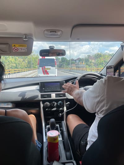 The image depicts the interior of a car, seen from the perspective of a passenger. The driver, wearing a white t-shirt and black shorts, is actively driving the car, with their hands on the steering wheel. A phone mounted on the windshield displays a navigation app. In the front passenger seat, a person is partially visible, wearing a black top. A red can of Pringles is placed in a cup holder between the seats. The car is on a road, with a bridge and surrounding vegetation visible through the windshield. In the distance, houses with traditional Balinese architecture can be seen, suggesting the location is likely Kerambitan, Indonesia. The sky is partly cloudy.