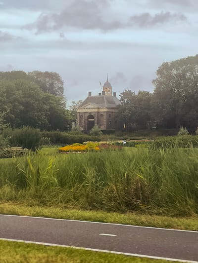 The image captures a frontal view of a historic building resembling a rotunda with a prominent cupola, situated amidst lush greenery and a flower garden. The building, constructed of stone with a classical architectural style, features a large arched entrance. It is surrounded by dense trees, and in the foreground, tall grass and a paved path with white markings are visible. The sky is overcast with visible clouds. The scene is identified as the entrance to the BnF (Bibliothèque nationale de France) in Enkhuizen, Netherlands. No people or explicit actions are depicted.