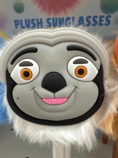 A close-up, eye-level, full shot features a plush sloth toy with large, cartoonish eyes and a wide smile. The sloth's face is gray with black accents around its eyes and nose, and a pink tongue is visible. Fluffy white fur surrounds the bottom of the toy. The background includes text that reads "PLUSH SUNGLASSES" in blue lettering and blurred colorful objects. The image was taken in Keila, Estonia.