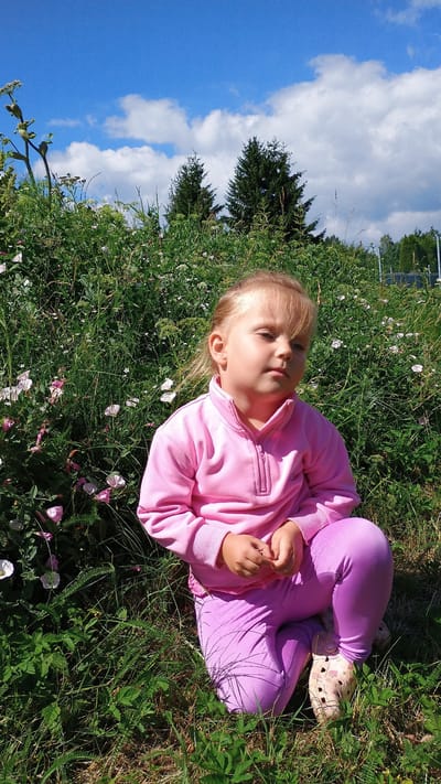 A young girl with blonde hair, wearing a pink zip-up sweatshirt and purple leggings, is crouching in a grassy area with blooming pink and white flowers. She is looking slightly down and to the right with a neutral expression. Behind her, a dense green bush forms a backdrop, with two dark green evergreen trees visible in the distance. The sky above is a clear blue with scattered white clouds. The scene appears to be an outdoor natural setting, possibly a park or a garden in Sokolov, Czechia.